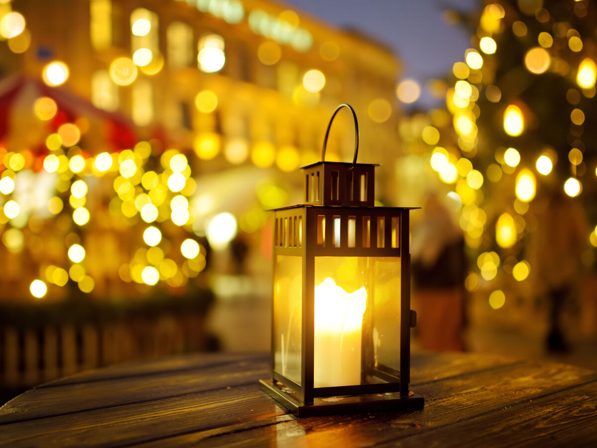 Laterne mit brennender Kerze steht auf einem Tisch. Im Hintergrund viele warme Lichtpunkte eines Weihnachtsmarktes.
