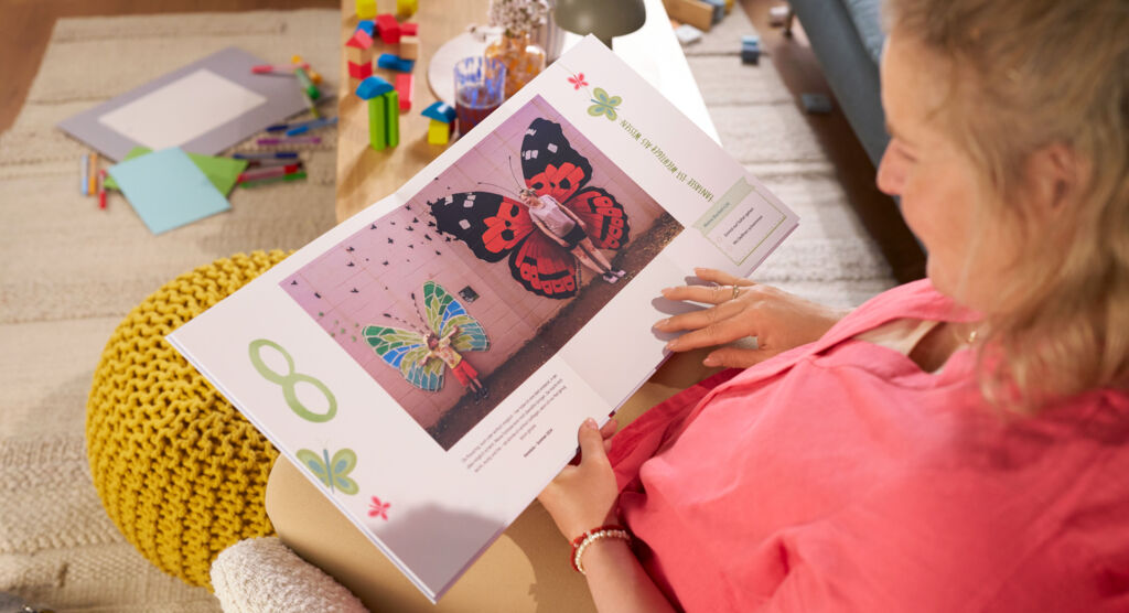 Eine Frau sitzt auf einem Sofa und blättert durch ein CEWE FOTOBUCH, die Doppelseite zeigt Fotomotive und die Zahl „8“.