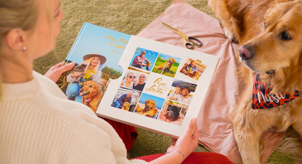 Eine Frau sitzt mit ihrem Hund auf einer Decke und betrachtet ihr bunt gestaltetes CEWE FOTOBUCH mit Schuber.