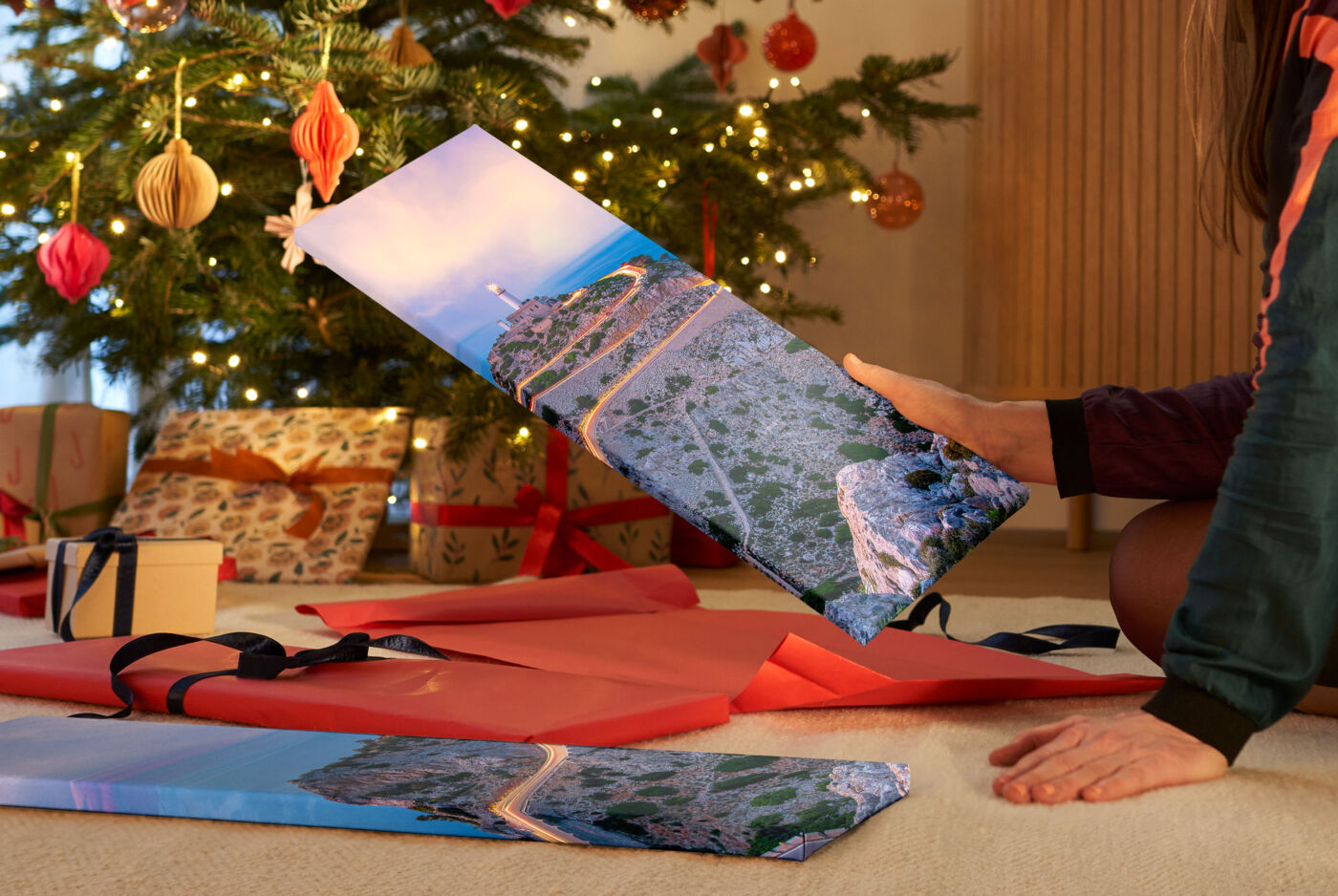 Das Foto zeigt ein Wohnzimmer mit festlich dekoriertem Tannenbaum. Davor sitzt eine Frau neben Geschenken auf dem Boden. In einer Hand hält