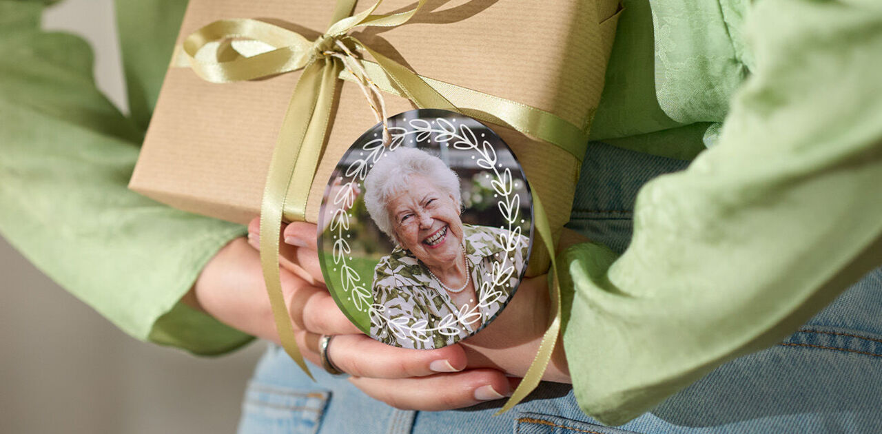 Person hält ein Geschenk mit Schleife und rundem Anhänger mit Foto einer lächelnden älteren Person hinter dem Rücken.