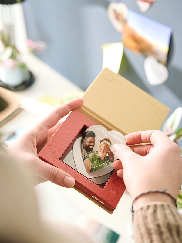 Hände einer Person halten eine geöffnete Geschenkbox, in der herzförmige Fotomagnete mit einem Paarfoto zu sehen sind.