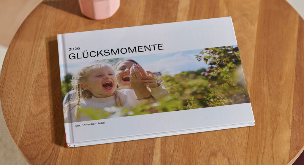 Ein Fotobuch liegt auf einem Holztisch. Auf dem Cover steht "Glücksmomente 2026" über einem Foto von zwei Mädchen im Grünen.