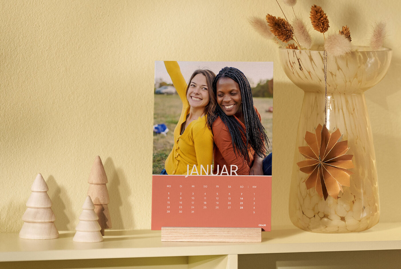 CEWE Fotokalender mit zwei Freundinnen im Freien, präsentiert in einem Holzaufsteller, dekoriert mit Holzfiguren und einer Vase mit Blumen